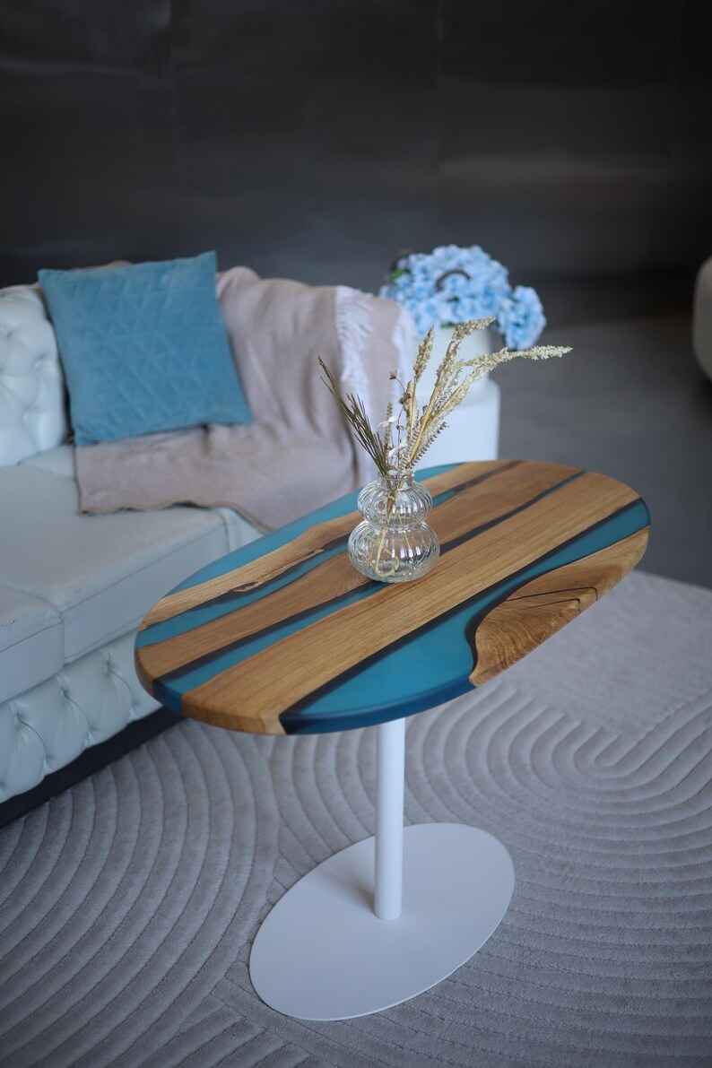 May include: An oval side table with a wooden top and blue resin river design. The table is supported by a white pedestal base. A glass vase with dried flowers sits on the table. The table is placed on a grey rug.