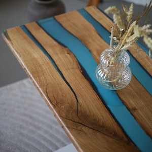May include: A handcrafted wooden table with a unique design. The table features natural wood planks with a vibrant blue resin river. A small glass vase with dried flowers sits on the table. The table's dimensions are approximately 36 inches long and 24 inches wide.