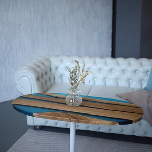 May include: Oval coffee table with a white base. The tabletop features a wood grain pattern with blue resin accents. A small glass vase with dried flowers sits on top. The table is approximately 91 cm long.