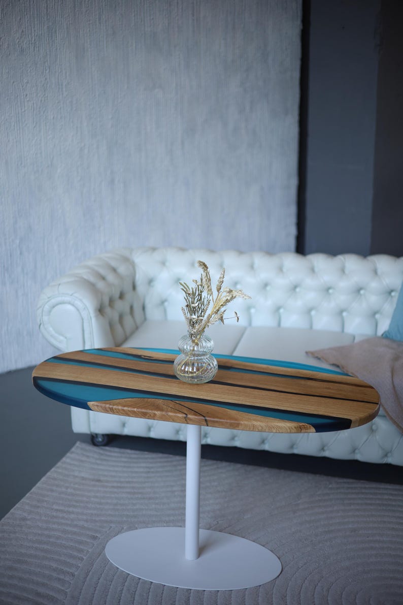 May include: Oval coffee table with a white base. The tabletop features a wood grain pattern with blue resin accents. A small glass vase with dried flowers sits on top. The table is approximately 91 cm long.