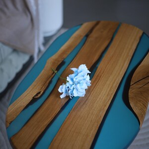 May include: Oval wooden table with teal resin detailing. The table has natural wood grain and a light blue flower. The table is supported by a white base. A decorative piece for a living room.