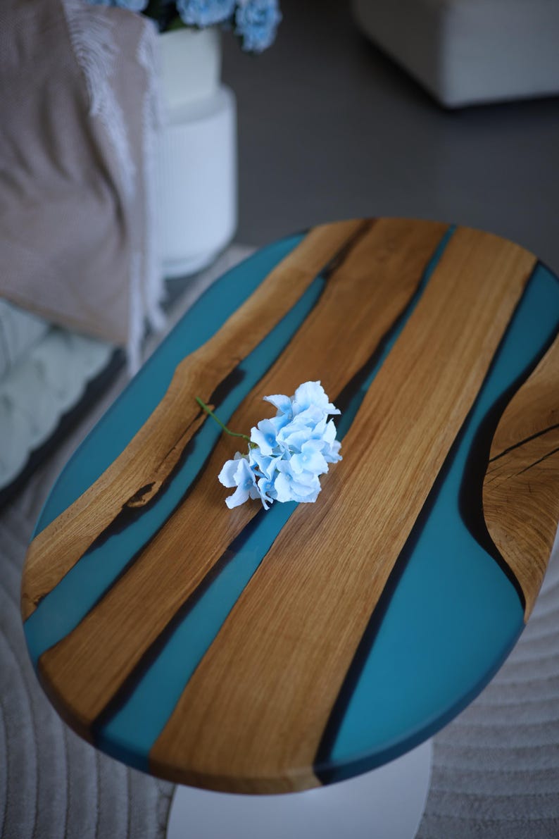 May include: Oval wooden table with teal resin detailing. The table has natural wood grain and a light blue flower. The table is supported by a white base. A decorative piece for a living room.