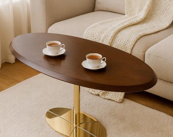 May include: A dark brown oval coffee table with a gold-coloured metal base. Two white teacups and saucers are on the table. The table is in a living room setting with a beige sofa and cream rug. The table is approximately 120 cm long.