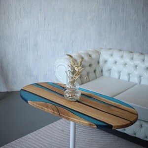 May include: Oval coffee table with a wooden top featuring blue resin and a clear glass vase with dried flowers. The table has a white metal base. The table is in front of a white tufted sofa.