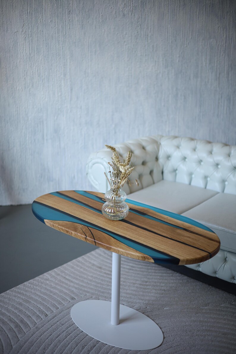 May include: Oval coffee table with a wooden top featuring blue resin and a clear glass vase with dried flowers. The table has a white metal base. The table is in front of a white tufted sofa.
