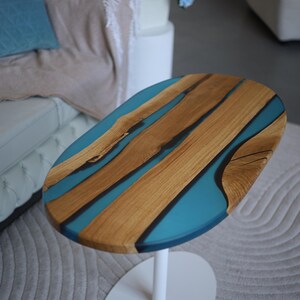 May include: Oval coffee table with a wooden top and turquoise resin inlays. The table has a white, single-pedestal base. The wood grain is visible. The table is set on a grey rug.