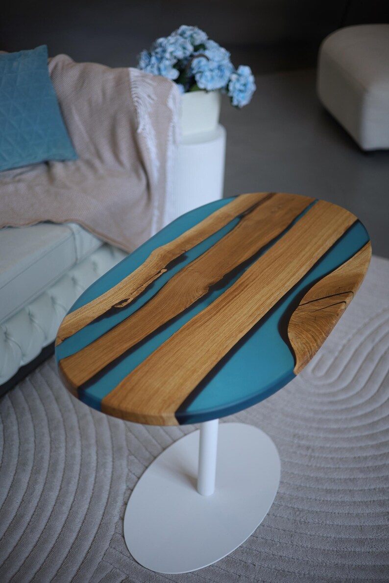 May include: Oval coffee table with a wooden top and turquoise resin inlays. The table has a white, single-pedestal base. The wood grain is visible. The table is set on a grey rug.
