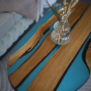 May include: Oval coffee table with a wooden top and teal resin detailing. A clear glass vase with dried flowers sits on the table. The table has a white base and is in a living room.