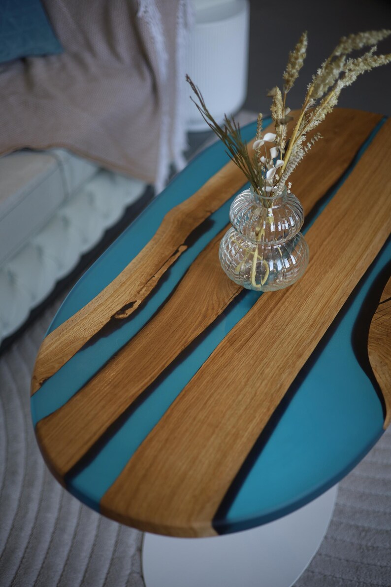 May include: Oval coffee table with a wooden top and teal resin detailing. A clear glass vase with dried flowers sits on the table. The table has a white base and is in a living room.