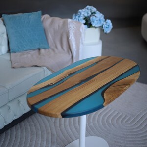 May include: Oval coffee table with a wooden top and teal resin detail. The tabletop has a natural wood grain and a glossy finish. The table has a white, single-pedestal base and is set on a neutral-coloured rug.