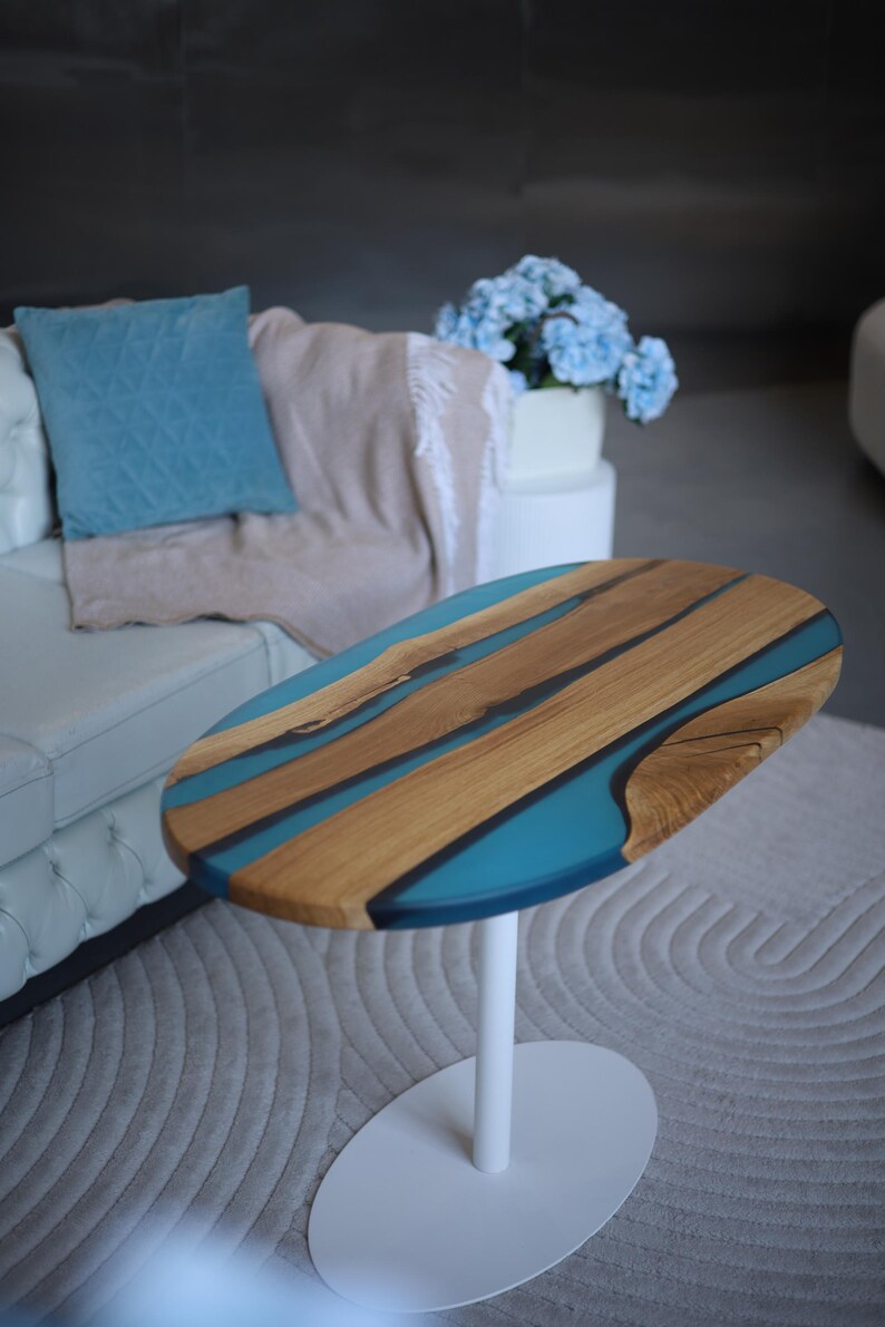 May include: Oval coffee table with a wooden top and teal resin detail. The tabletop has a natural wood grain and a glossy finish. The table has a white, single-pedestal base and is set on a neutral-coloured rug.