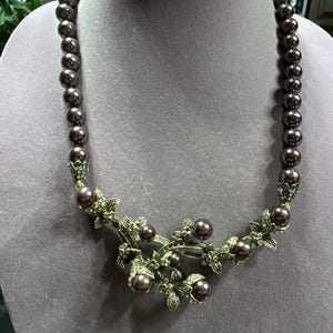 Könnte beinhalten: Eine braune Perlenkette mit floralem Design. Die Halskette hat dunkelbraune Perlen und einen dekorativen bronzefarbenen Blumenanhänger mit grünen Akzenten und dunkelbraunen perlenartigen Perlen.