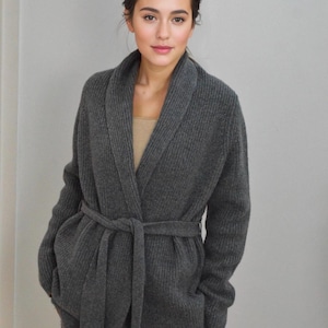 May include: A dark grey knitted cardigan with a shawl collar and a matching belt tied at the waist. The cardigan is part of a set and appears to be made of a soft, warm material.