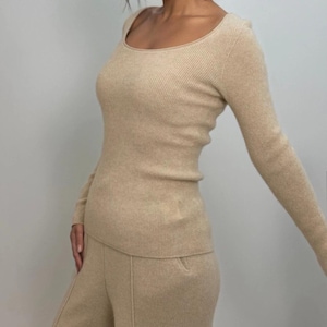 Può includere: Un top in maglia a coste beige con scollo tondo e maniche lunghe, abbinato a pantaloni a gamba larga coordinati. L'outfit è di colore neutro, creando un look coerente e minimalista. Il tessuto appare morbido e confortevole.