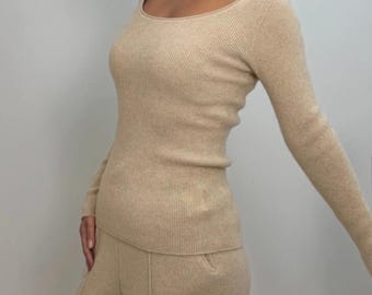 May include: A beige ribbed knit top with a scoop neckline and long sleeves, paired with matching wide-leg pants. The outfit is a neutral color, creating a cohesive and minimalist look. The fabric appears soft and comfortable.