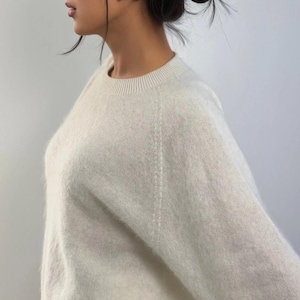 Może przedstawiać: Kremowy sweter z okrągłym dekoltem i długimi rękawami. Sweter ma miękki, teksturowany wygląd i prążkowany dekolt. Modelka ma ciemne włosy spięte w kok i nosi małe kolczyki.