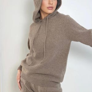 May include: A taupe-coloured long-sleeved hooded jumper featuring a ribbed knit texture and matching drawstring cords. The garment includes a hood and a straight hemline, crafted from a soft-looking knit fabric in a neutral earth tone.