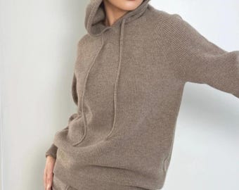 May include: A taupe-colored long-sleeved hooded sweater featuring a ribbed knit texture and matching drawstring cords. The garment includes a hood and a straight hemline, crafted from a soft-looking knit fabric in a neutral earth tone.