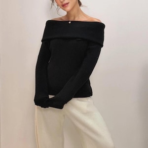 Può includere: Un maglione nero a maniche lunghe caratterizzato da un ampio scollo ripiegato che lascia le spalle scoperte e una trama a coste, abbinato a pantaloni larghi color bianco sporco con taglio dritto e finitura in tessuto liscio.
