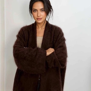 May include: A dark brown, fuzzy, open-front cardigan with long, wide sleeves. The garment features a soft, textured knit construction with a relaxed fit, worn over a simple, light-coloured, ribbed tank top underneath the dark brown knit layer.