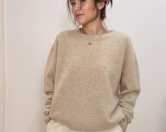 May include: A beige crew neck sweater with long sleeves. The sweater is worn with light-coloured trousers. The model is wearing a necklace. The sweater appears to be made of a soft, knitted material.