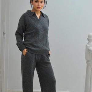 Op de afbeelding: Een antracietgrijze gebreide set, bestaande uit een top met lange mouwen en kraag en een bijpassende broek met wijde pijpen. De outfit is gemaakt van een zacht, getextureerd materiaal. Het model staat op een trap.