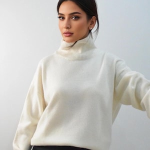 Puede incluir: Un suéter de cuello alto color crema con mangas largas. El suéter se combina con una falda negra. La modelo lleva un pendiente de botón. El fondo es una pared blanca lisa.