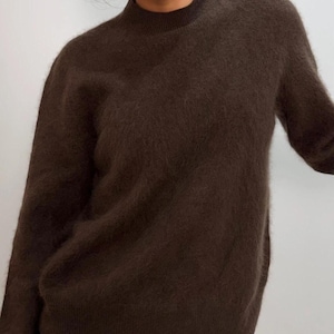 Puede incluir: Un suéter de cuello alto marrón oscuro. El suéter tiene una textura suave y difusa y mangas largas. El escote es un cuello alto y ajustado. El suéter es de color liso y parece estar hecho de un material cálido.