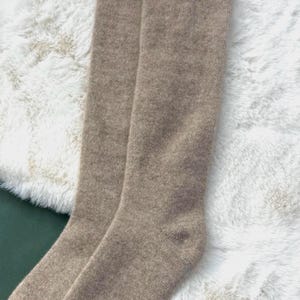 May include: A pair of knee-high socks in a neutral tan color. The socks are made of a soft, knitted material and are displayed on a white, fluffy surface. The socks are folded neatly, showcasing their length and texture.