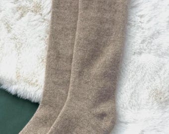 May include: A pair of knee-high socks in a neutral tan color. The socks are made of a soft, knitted material and are displayed on a white, fluffy surface. The socks are folded neatly, showcasing their length and texture.