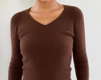May include: A brown, long-sleeved sweater with a V-neck. The ribbed knit top is a solid color and appears to be made of a soft material. The model is wearing the sweater.