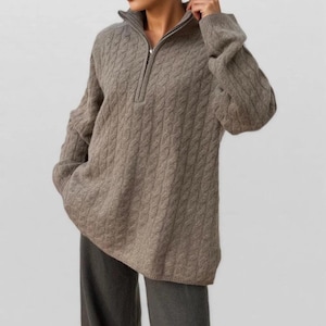 May include: A taupe cable-knit sweater with a quarter-zip neckline. The sweater has a relaxed fit and long sleeves. The model is wearing gray pants. This sweater is perfect for fall and winter.