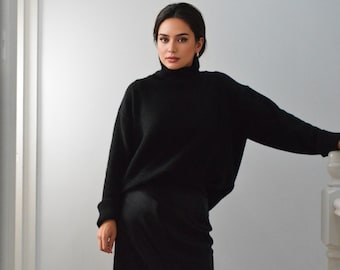 May include: A woman wearing a black turtleneck sweater and a long black skirt. The skirt has a lace trim at the bottom. The woman is leaning against a white wall and a white banister.