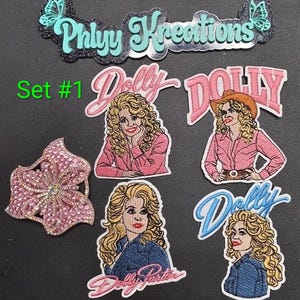 May include: A collection of embroidered patches including a logo reading Phlyy Kreations with butterflies, a pink flower patch, and four patches featuring a blonde woman with the text Dolly or Dolly Parton in pink and blue script lettering.