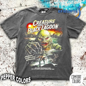 May include: Dark gray t-shirt featuring a vintage movie poster design for "Creature from the Black Lagoon." The graphic includes the creature, text, and a sunset background. The shirt has the words "Amazing! Startling! Shocking!" printed on it.