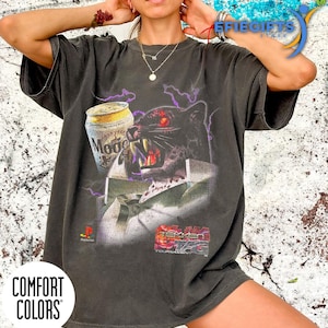 Puede incluir: Una taza de cerámica blanca con un asa negra y el texto "But First, Coffee" en negrita negra. La taza se encuentra sobre un fondo borroso, sugiriendo un ambiente acogedor.