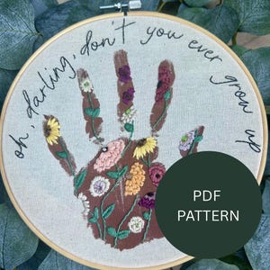 Könnte beinhalten: Stickrahmen-Kunst mit einem braunen Handabdruck-Design, gefüllt mit bunten gestickten Blumen. Der Text "oh darling, don't you ever grow up" ist am Rand gestickt. Ein dunkelgrüner Kreis mit "PDF PATTERN" ist im Bild.