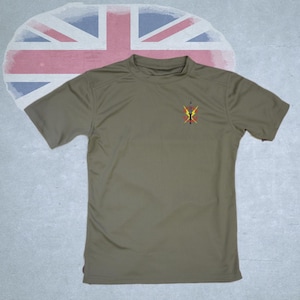 May include: Olive green short-sleeve t-shirt with a small emblem on the left chest. The emblem features a yellow, red, and black design. The background includes a faded Union Jack flag.