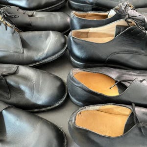 May include: A collection of black leather dress shoes with laces. The shoes are arranged closely together, showcasing their classic design and polished appearance. Some shoes have visible insoles.