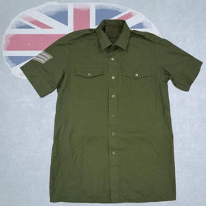 Può includere: Una camicia a maniche corte color verde oliva con colletto a punta, due tasche sul petto con patta abbottonata e spalline. Sulla manica destra è visibile una toppa rettangolare con tre strisce orizzontali di colore chiaro.