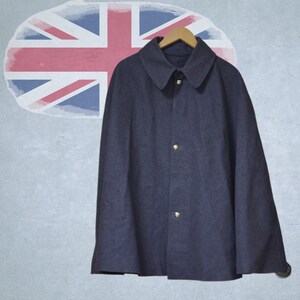 Puede incluir: Una capa azul marino con cuello y tres botones dorados, colgada de una percha de madera. El fondo presenta un diseño descolorido de la bandera de la Union Jack en una pared gris.