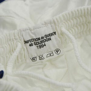 May include: White clothing label with black text that reads "Confection du Quercy 46 Gourdon 1994". The label also has a black and white washing instruction symbol.