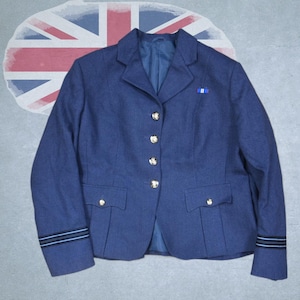 Puede incluir: Una blazer azul marino con botones dorados y dos bolsillos delanteros. La blazer tiene una cinta azul en la solapa y rayas en las mangas. El fondo presenta una bandera de la Union Jack descolorida.