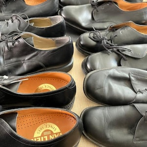 May include: A collection of black leather dress shoes with laces. The shoes have a classic design with a rounded toe and a low heel. The insoles are visible, featuring a gold-colored stamp. The shoes are arranged in a close-up shot.
