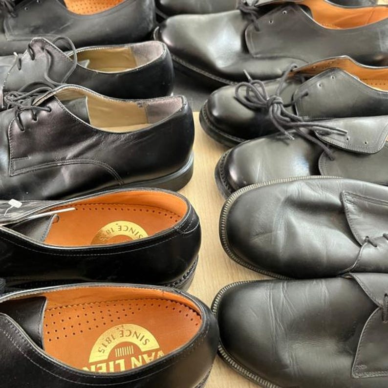 May include: A collection of black leather dress shoes with laces. The shoes have a classic design with a rounded toe and a low heel. The insoles are visible, featuring a gold-colored stamp. The shoes are arranged in a close-up shot.