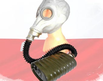 May include: A vintage gas mask with a white facepiece, circular eyepieces, and a black corrugated hose connected to a green canister. The mask is displayed on a mannequin head against a red and white background.