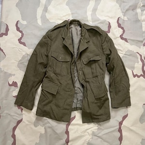 Peut inclure: Veste militaire vert olive avec plusieurs poches avant et fermetures boutonnées. La veste a un col et des épaulettes. La doublure est d'une nuance de vert plus claire. La veste est présentée sur un fond à motif de camouflage.