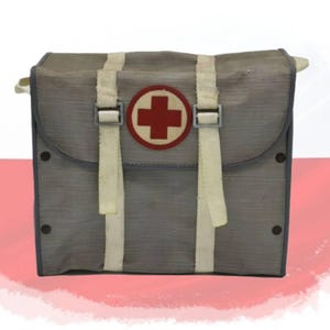 Op de afbeelding: Een grijze rechthoekige tas met een opvallende ronde patch met een rood kruis op een witte achtergrond. De tas is voorzien van twee verticale, gebroken witte stoffen banden met verstelbare gespen, donkergrijze randen en vier donkere sluitingen.