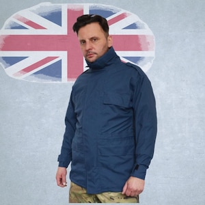 以下が含まれることがあります： ネイビーブルーのジャケット。複数のポケットとハイカラーが特徴。迷彩柄のパンツを着用した人物が写っています。背景には、色あせたユニオンジャックの旗があります。