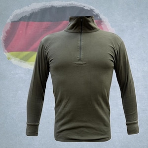 Könnte beinhalten: Olivgrünes Langarmshirt mit Stehkragen und einem Viertel-Reißverschluss. Das Shirt hat gerippte Bündchen und ein schlichtes, klassisches Design. Im Hintergrund ist eine verblasste deutsche Flagge zu sehen.
